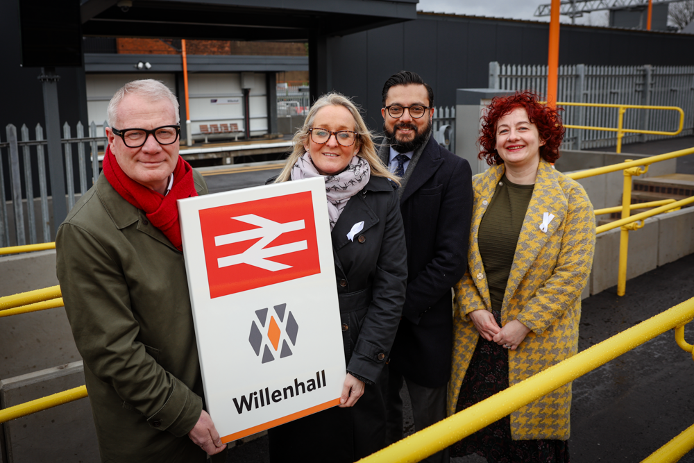Four people standing in front of a railway station entrance - two are holding a sign with a British rail logo and Willenhall on it.